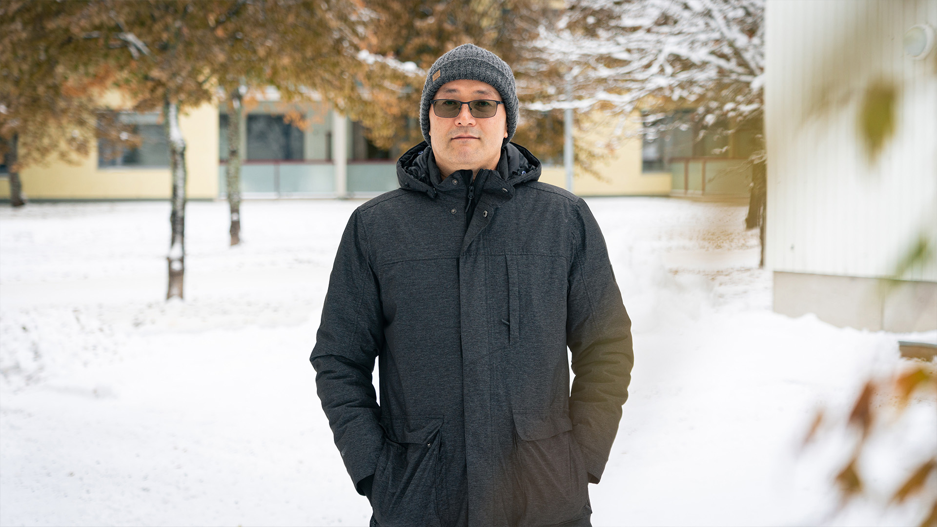 Mies seisoo ulkona katse kameraan päin. Taustalla luminen piha ja asutusta.