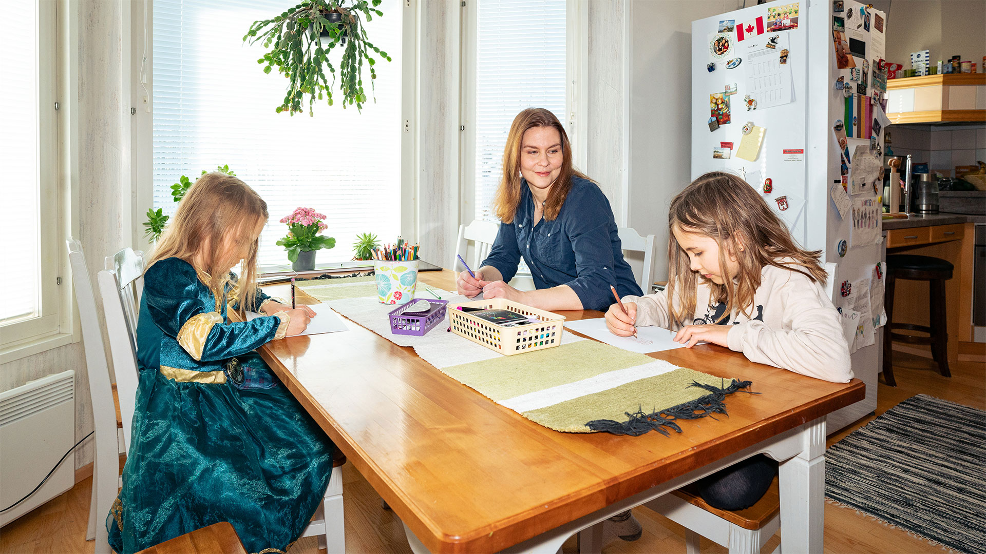 Nainen ja lapset istuvat ruokapöydän äärellä piirtämässä.