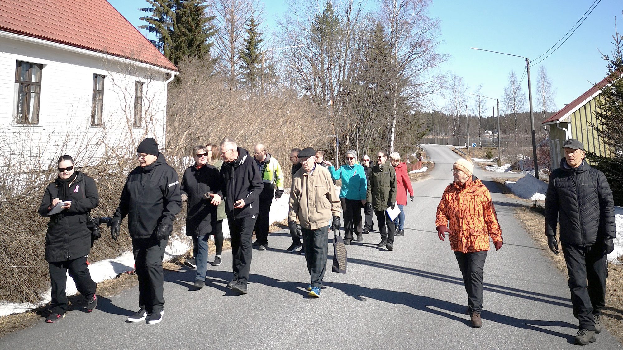 Joukko ihmisiä kävelemässä ja keskustelemassa maaseutumaisella asuinalueella.