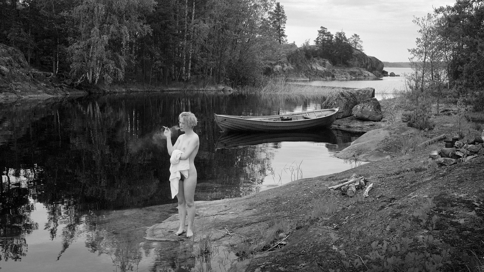 Mustavalkoinen valokuva, jossa nainen seisoo alastoman pyyhkeen kanssa rannalla tupakoimassa.