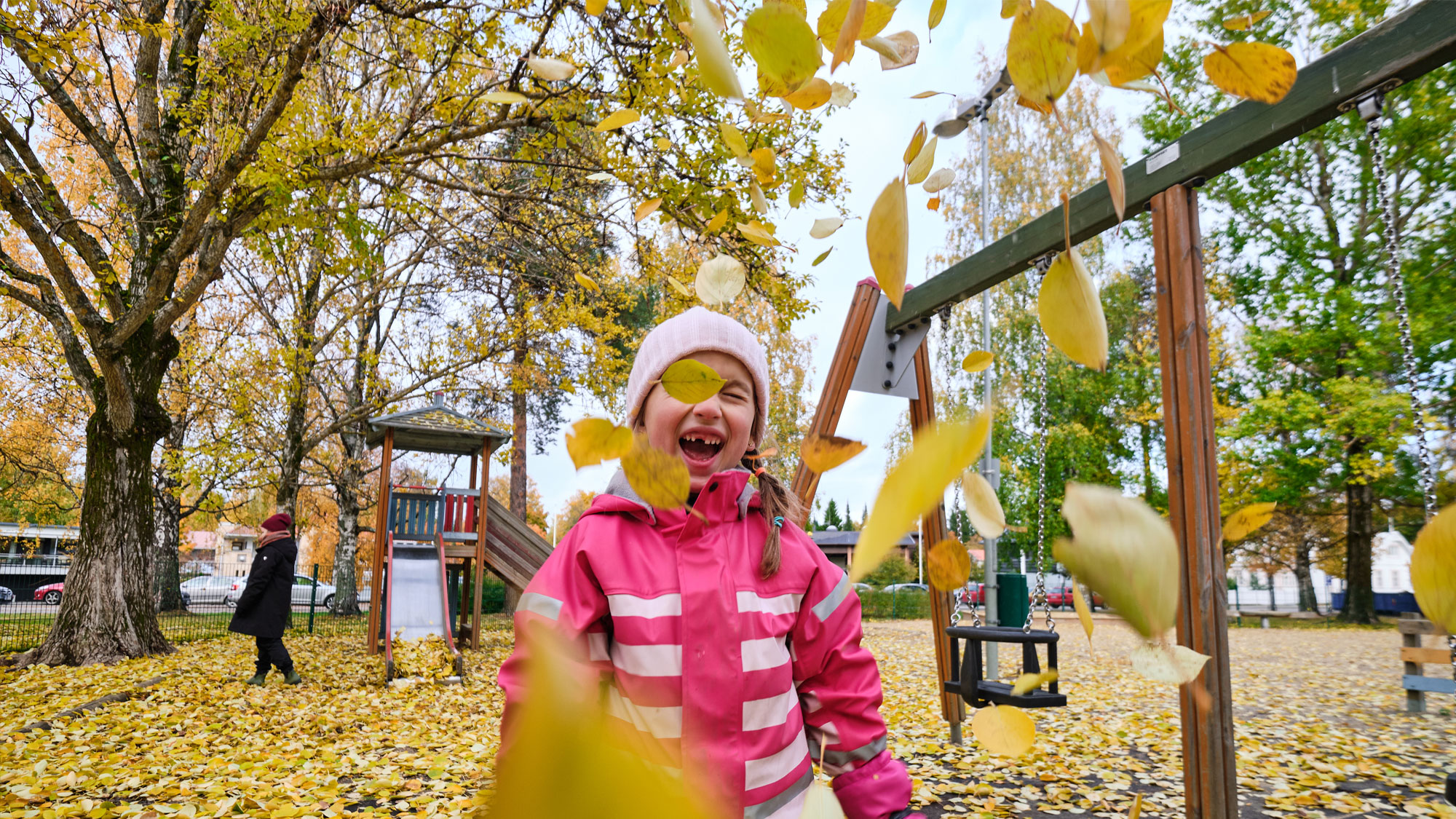 Syksyllä puistossa leikkivä lapsi heittää ilmaan puista pudonneita lehtiä.