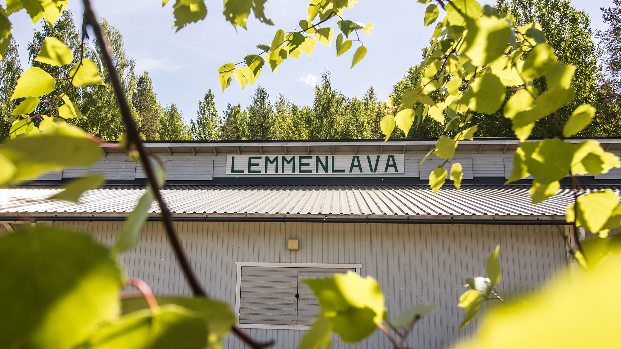 Puuverhoiltu vaalean sävyinen rakennus, jonka katonrajassa on kyltti Lemmenlava. Rakennuksen edustalla on koivuja.