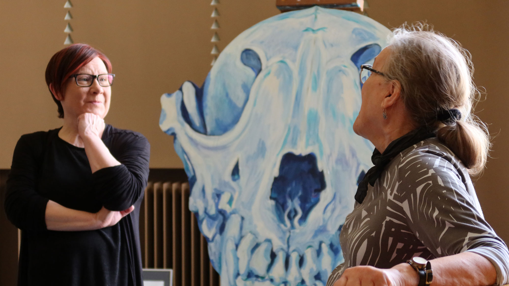 Teatterinjohtaja Iiristiina Varilo ja kuvataiteilija Arja Valkonen-Goldblatt keskustelevat teatterin aulassa. Taustalla sinisävyinen maalaus.
